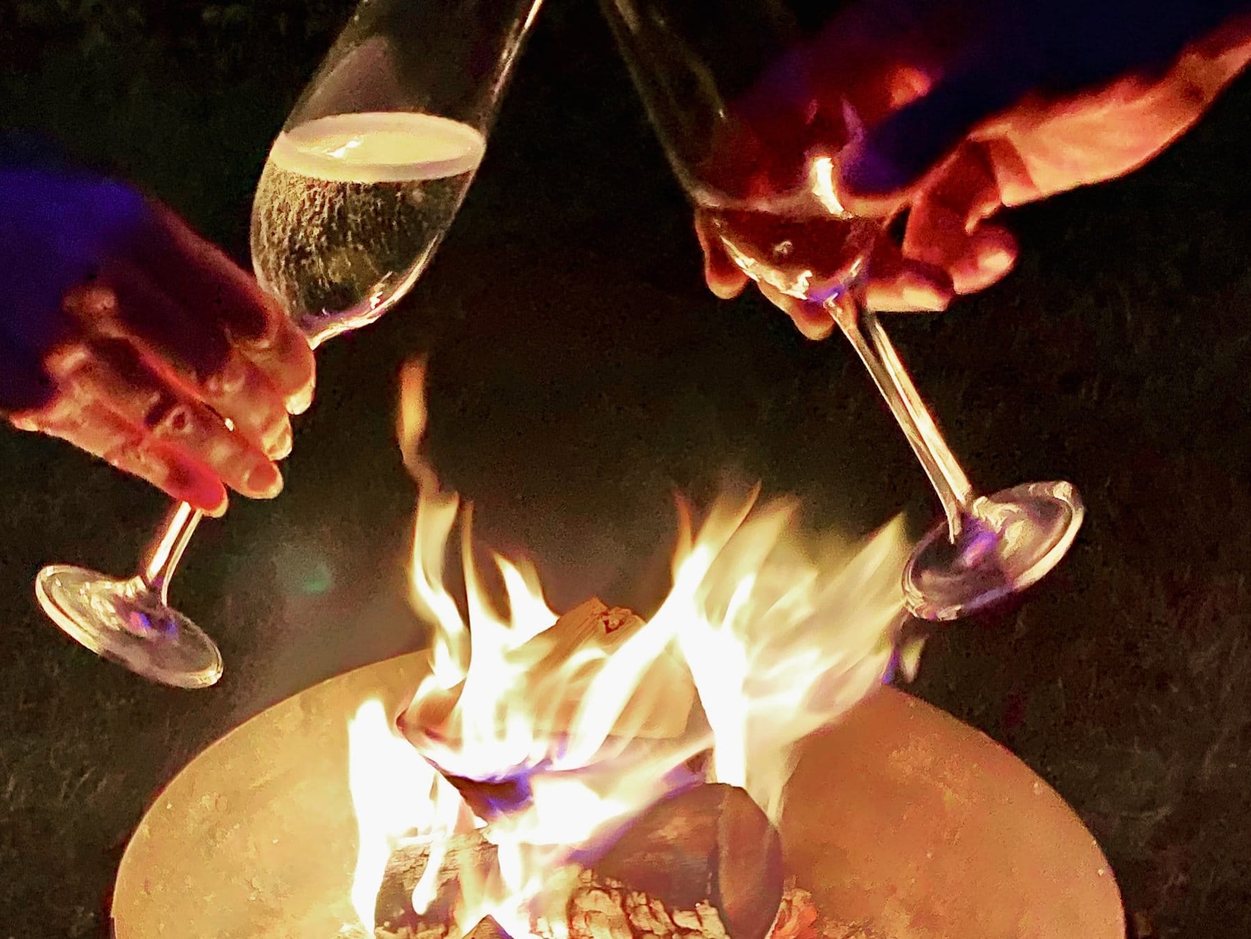 Guests holding glasses of champagne by a glowing rusty metal firebowl on a quiet evening at Batcombe Vale campsite in Somerset.