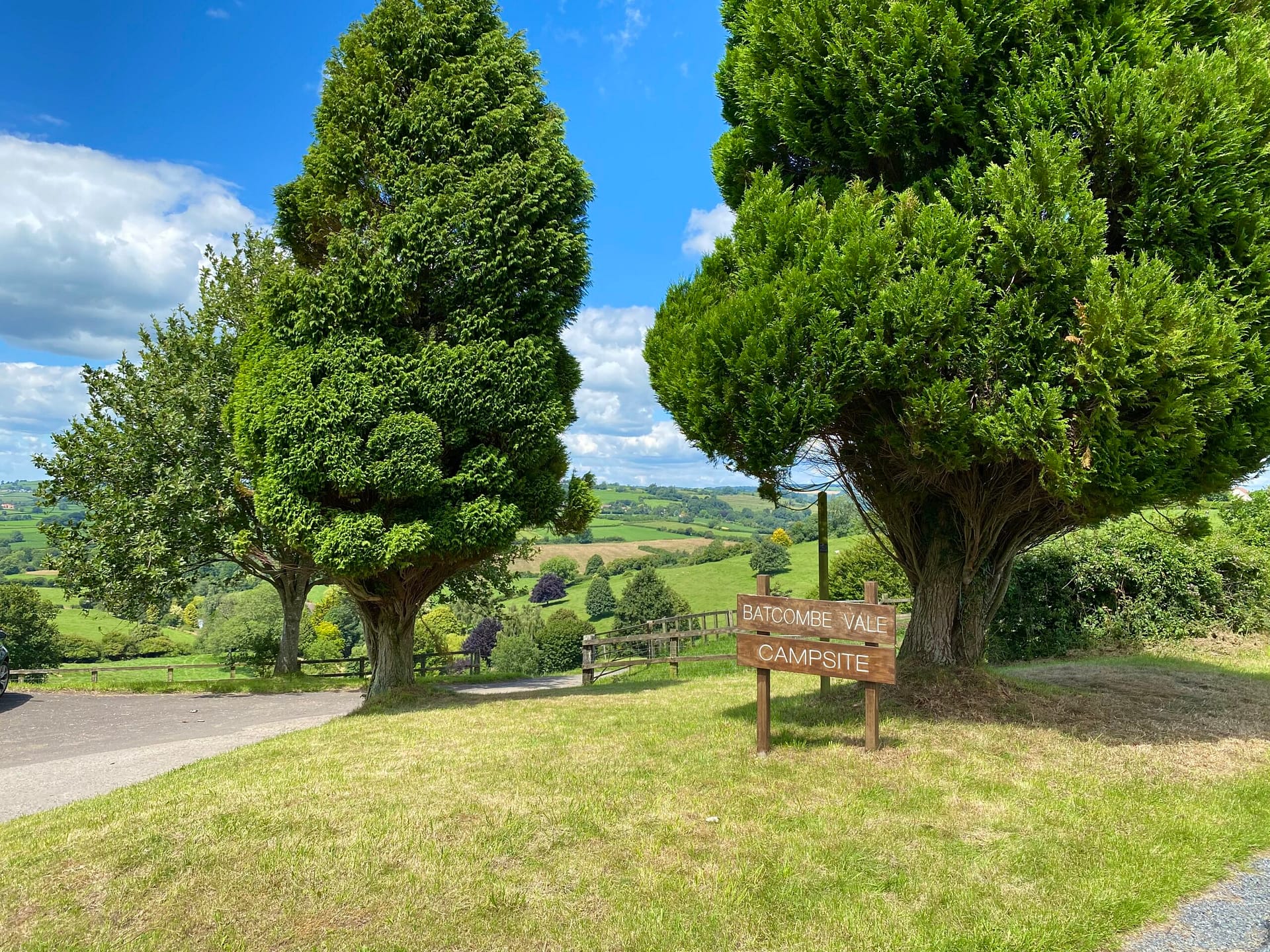 Batcombe Vale campsite entrance sign beneath sculpted topiary trees in Somerset, with a winding path and rolling green hills in the background on a sunny summer's day.