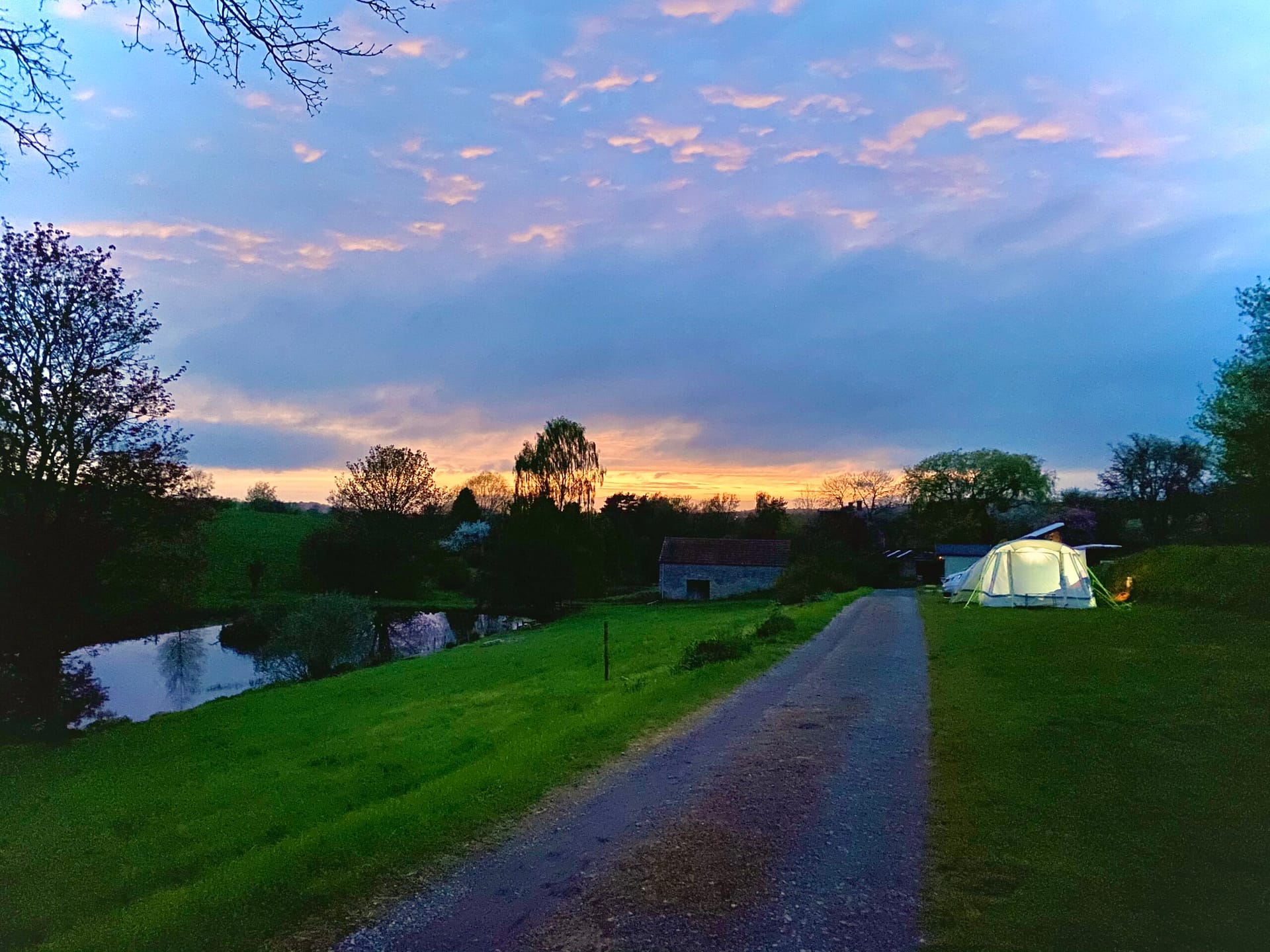 An illuminated, glowing geodesic dome glamping tent positioned on a grassy bank at dusk, overlooking a peaceful lake and the Somerset valley skyline at Batcombe Vale adults-only campsite.