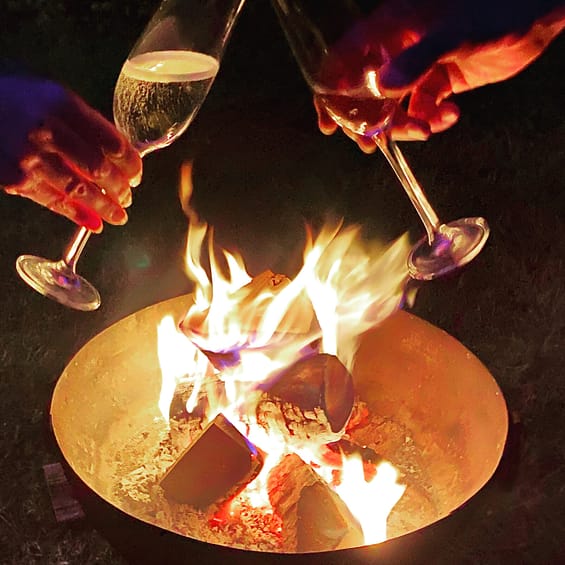 two people's hands holding a glass of champagne each over a kadai firebowl with burning logs at night